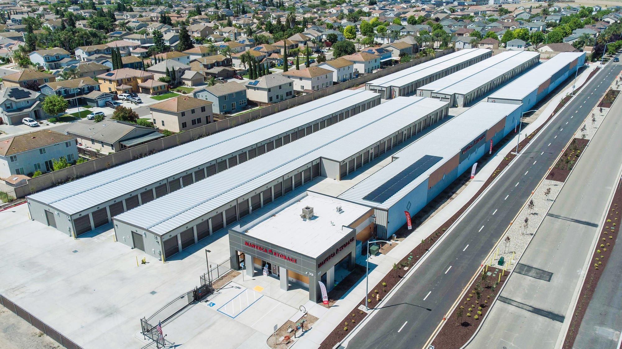 Aerial view of Manteca Storage: four long rows of drive-up units and a solar-panelled building beside a Manteca neighborhood